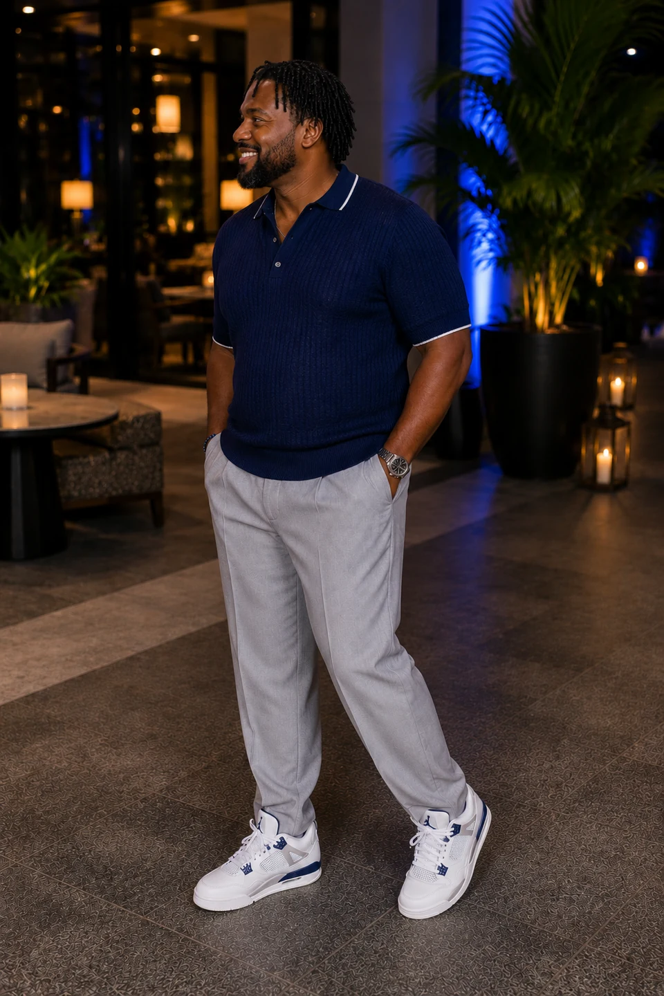 Dark-skinned Black man with short locs in a textured navy knit polo, light-gray tailored trousers, and white basketball-style sneakers.